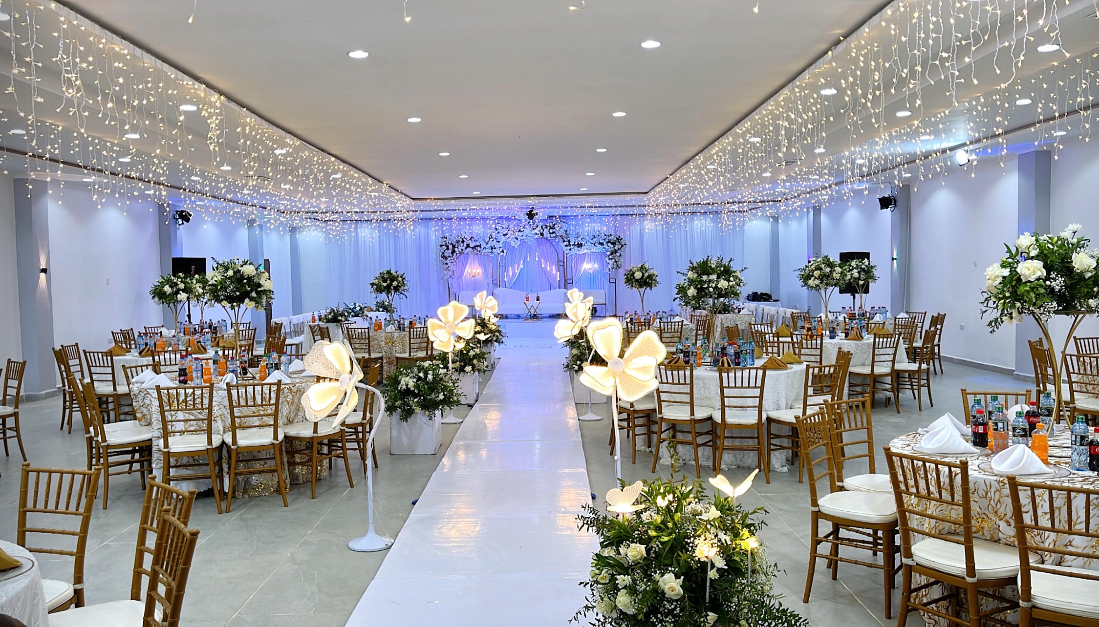Hall with curtains of fairy lights, gold chairs and a white aisle lined with glowing flowers