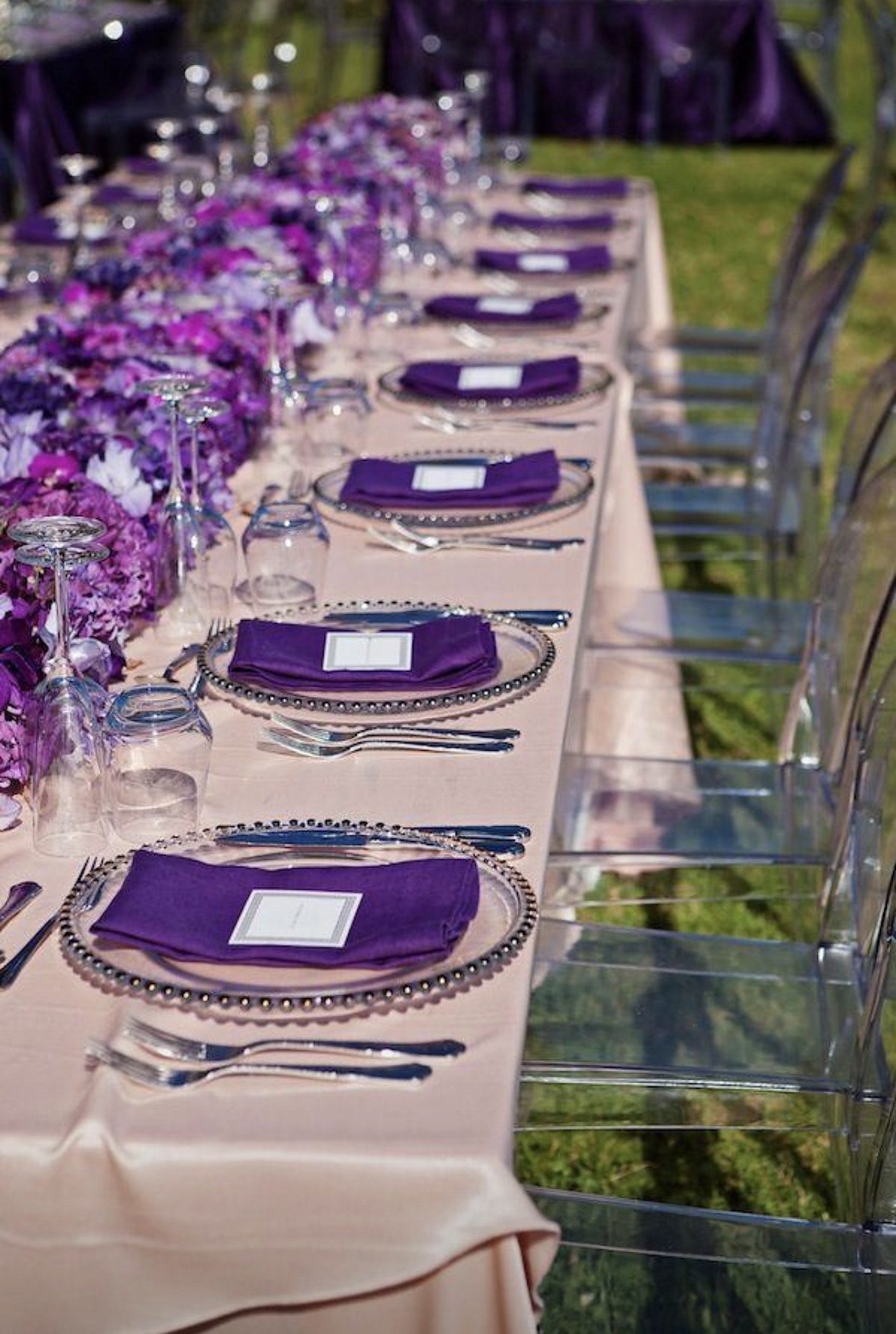 Purple napkins on beaded chargers along a long table with clear chairs