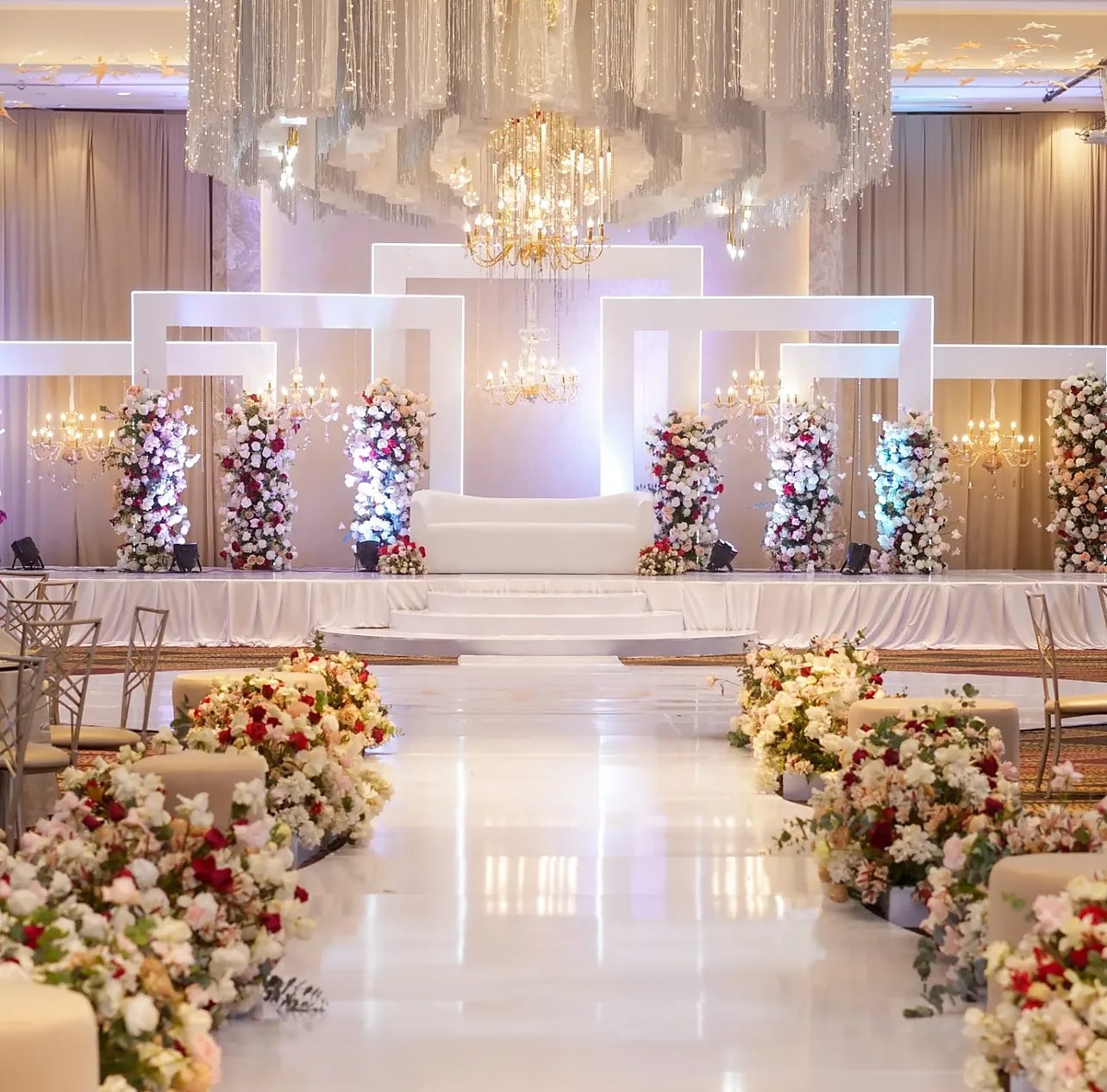 White stage with floral columns and an ivory sofa at the end of a rose-lined aisle