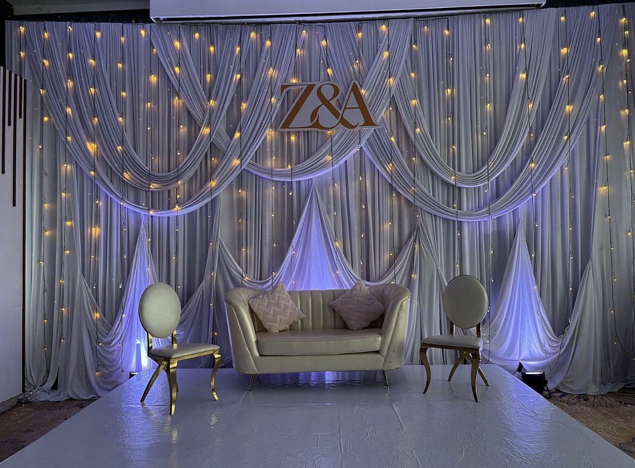 Stage of fairy-lit white drapery with a gold monogram and an ivory loveseat