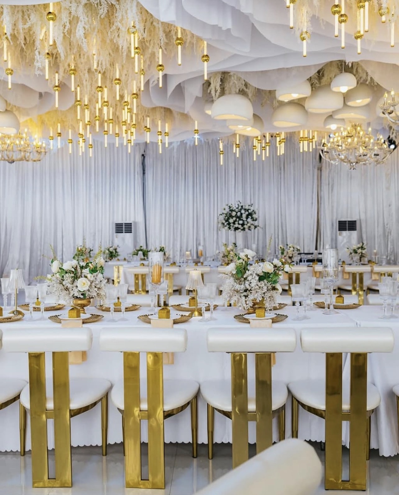 Long tables with gold-framed chairs and white florals under suspended candles