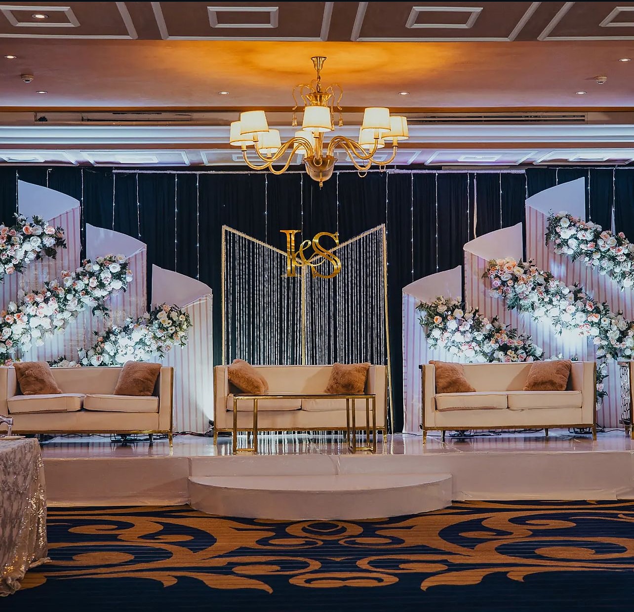 Ballroom stage with white floral panels, a gold monogram and three tan sofas