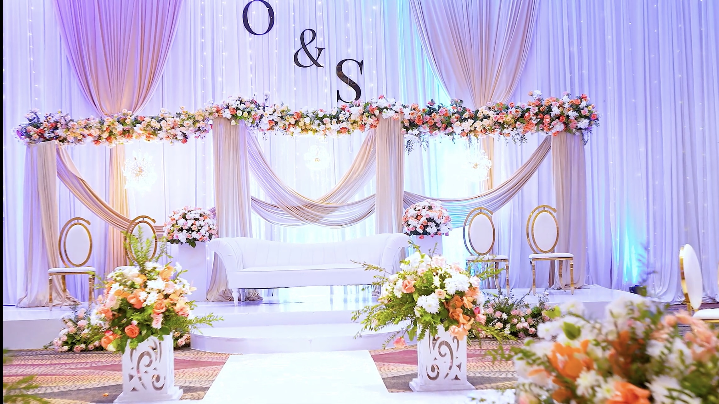 Wedding stage with draped columns, a garland of peach and white roses, gold chairs and a monogram above an ivory sofa