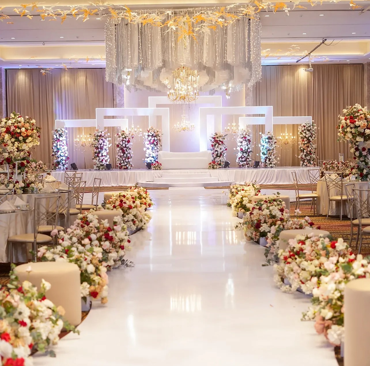 White aisle lined with red and ivory roses leading to a white stage under crystal chandeliers